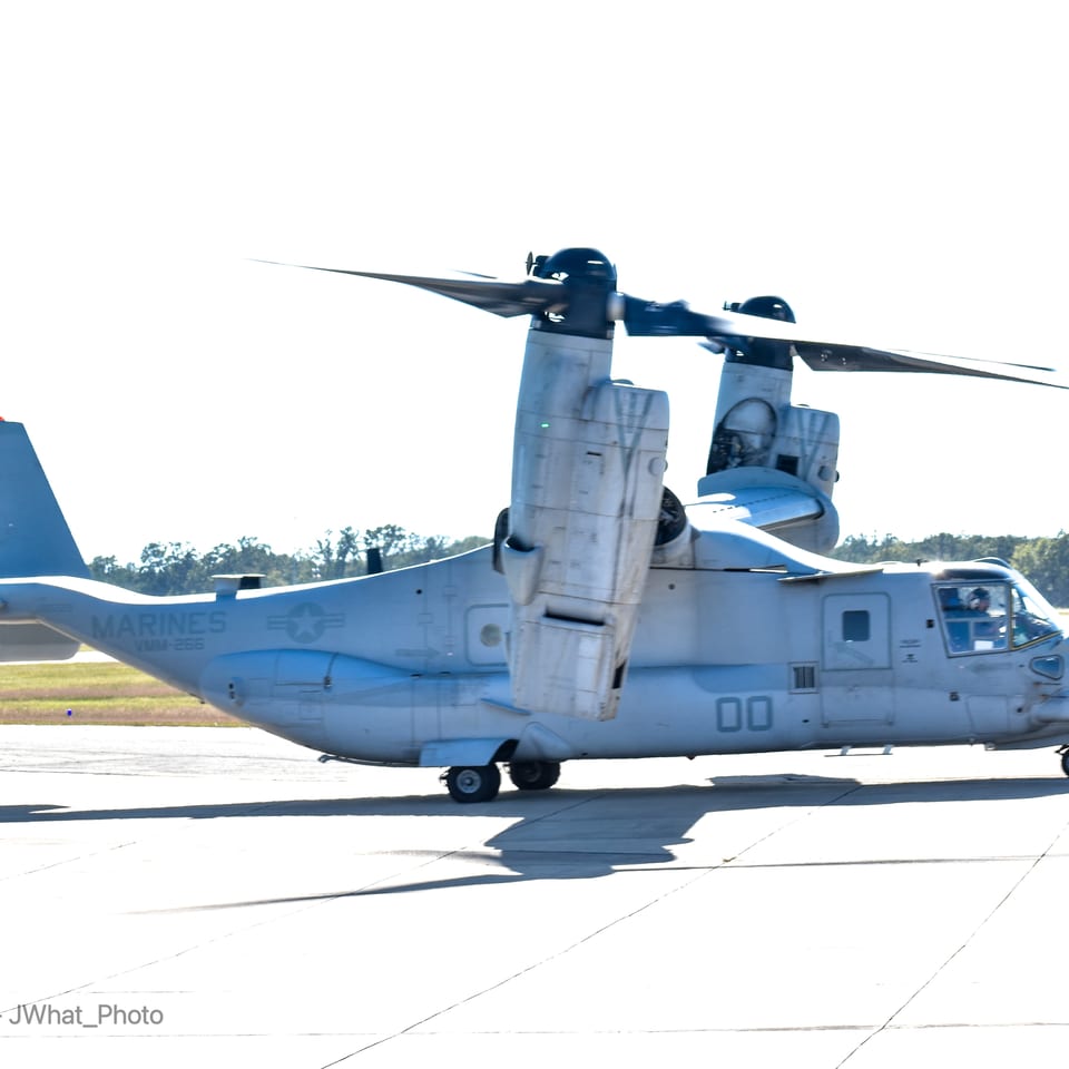 A U.S. Marine Corps tiltrotor aircraft sits on a runway with its rotors extended and grassy airfield behind it.