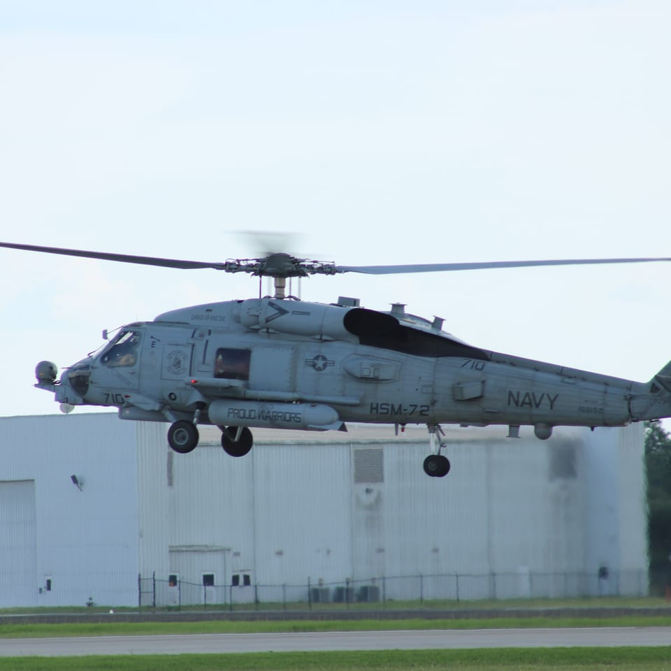 Navy military helicopter hovering above a runway near an industrial hangar, with landing gear lowered and rotors spinning.
