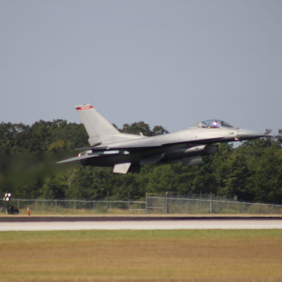 A fighter jet flies low over a runway, with trees, fencing, and grassy fields in the background.