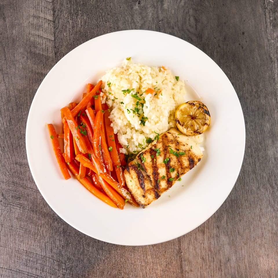 Grilled fish with rice, glazed carrots, and a charred lemon on a white plate.