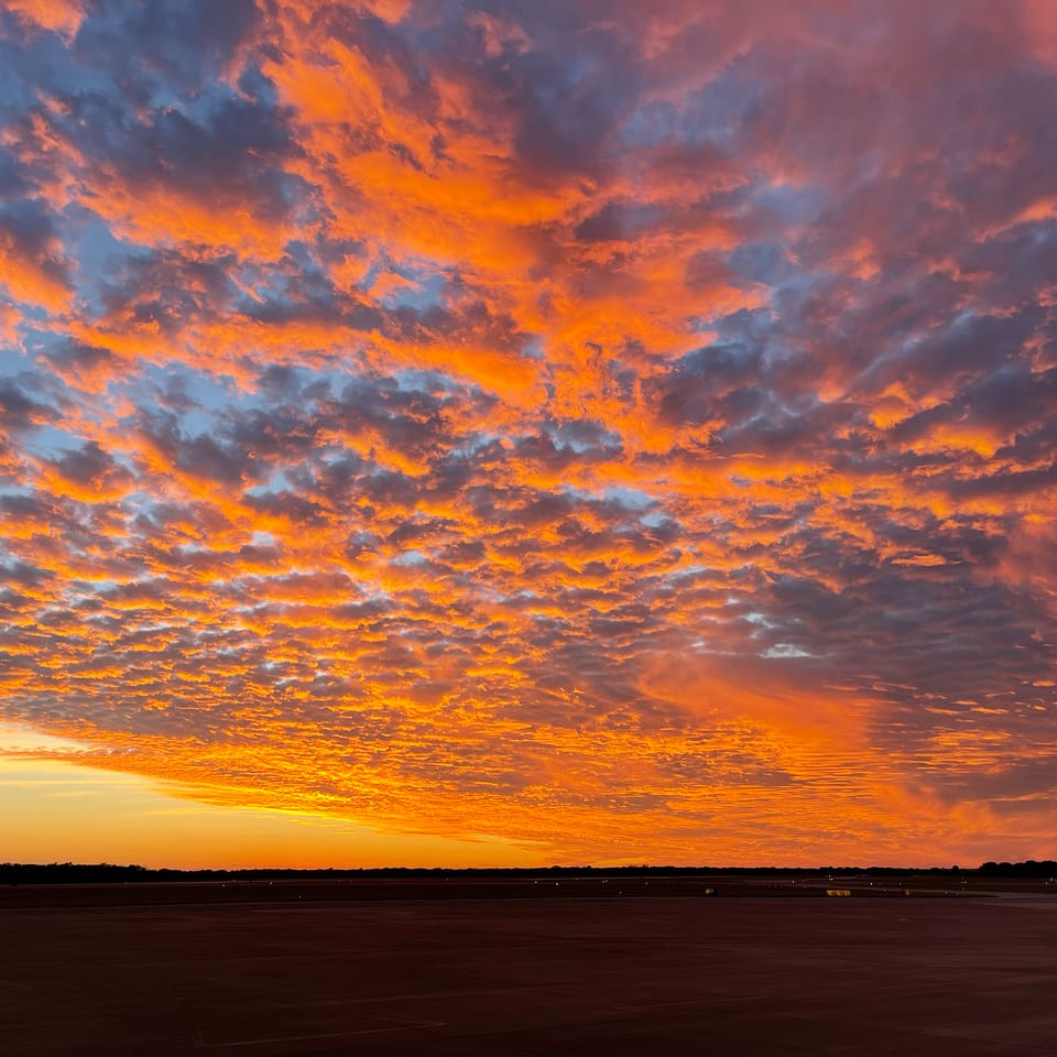 Vivid orange and purple sunset clouds spread above a dark airport runway and distant flat horizon.