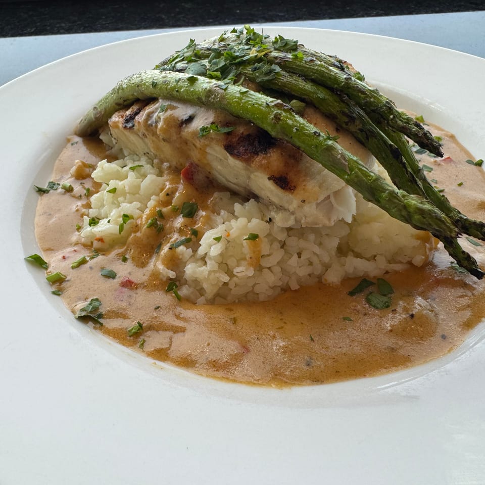 Grilled fish over white rice with roasted asparagus, chopped herbs, and creamy sauce on a white plate.