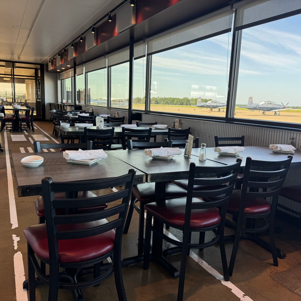 Restaurant dining room with set tables and red-cushioned chairs overlooking several parked aircraft through large windows.