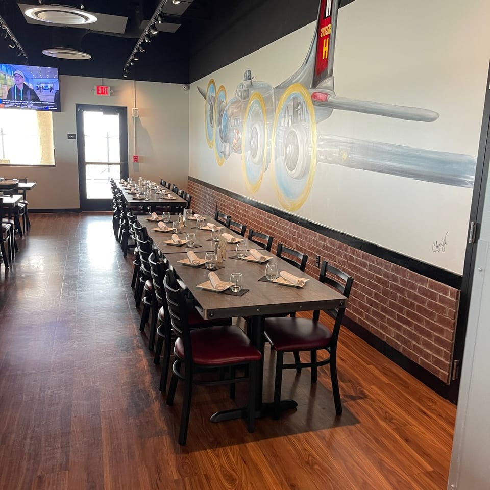 Restaurant dining room with neatly set tables, brick-accented walls, hardwood floors, and a colorful vintage airplane mural.