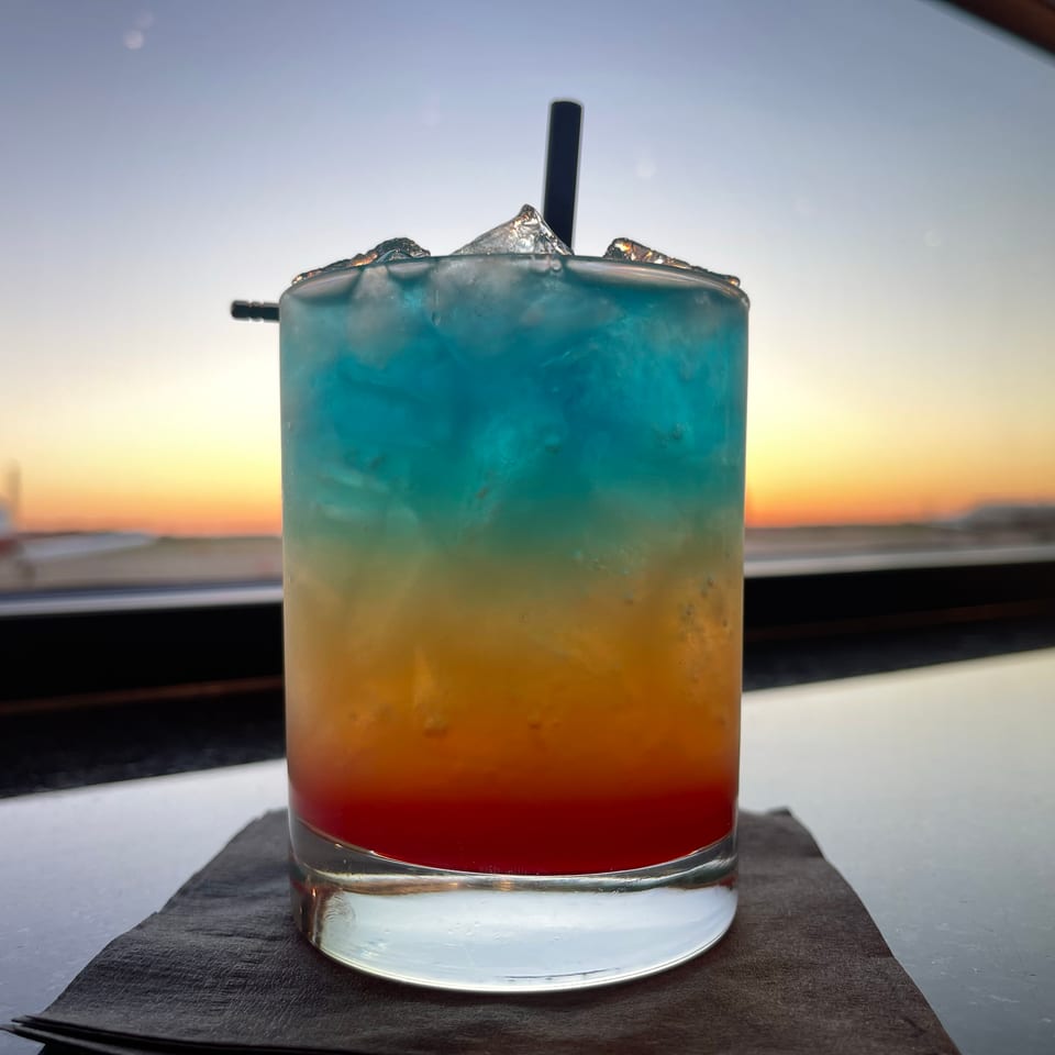 Layered blue, yellow, and red cocktail with ice and straw on a napkin, silhouetted against a sunset view.