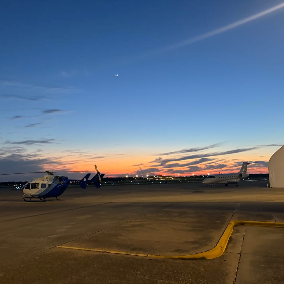 Helicopter and private jet parked on an airport tarmac beneath a sunset sky, with a large hangar visible on the right.