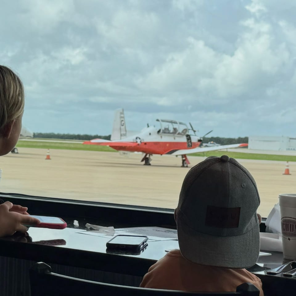 Family watches a red-and-white vintage airplane from a restaurant window overlooking an airport runway.