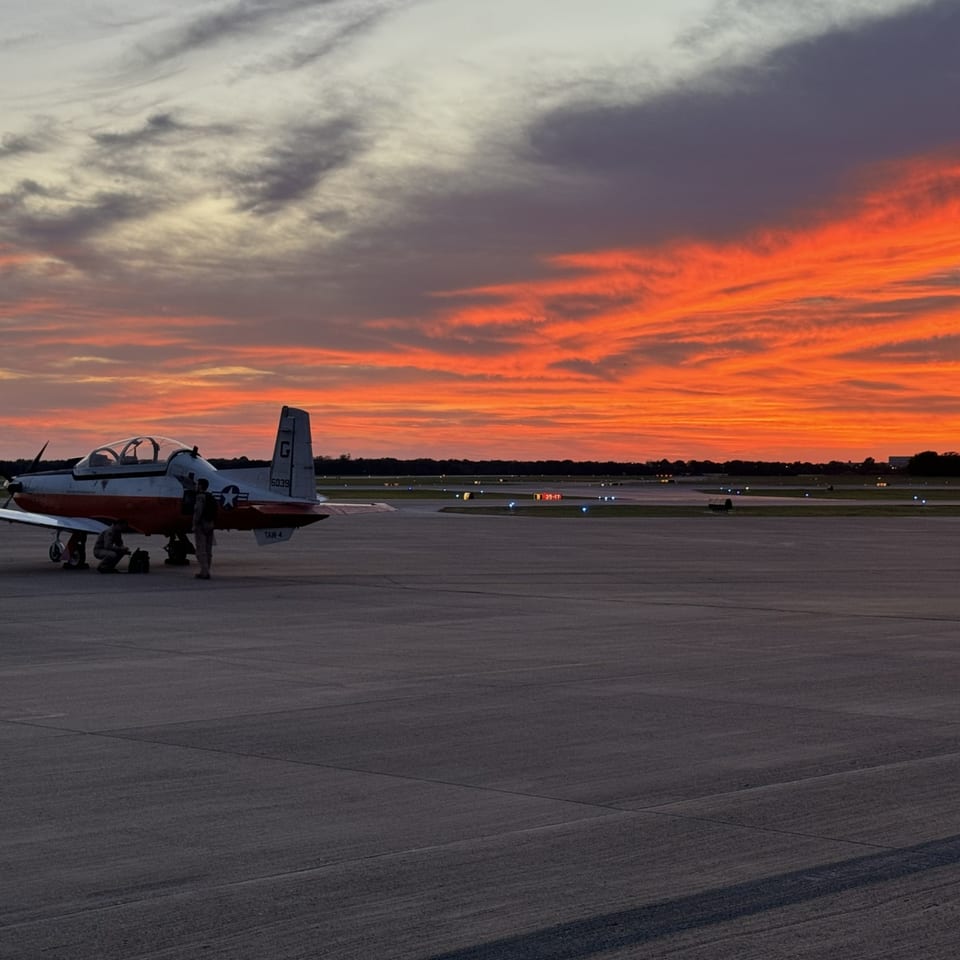 A training aircraft parked on the runway under a fiery red and orange sunset.