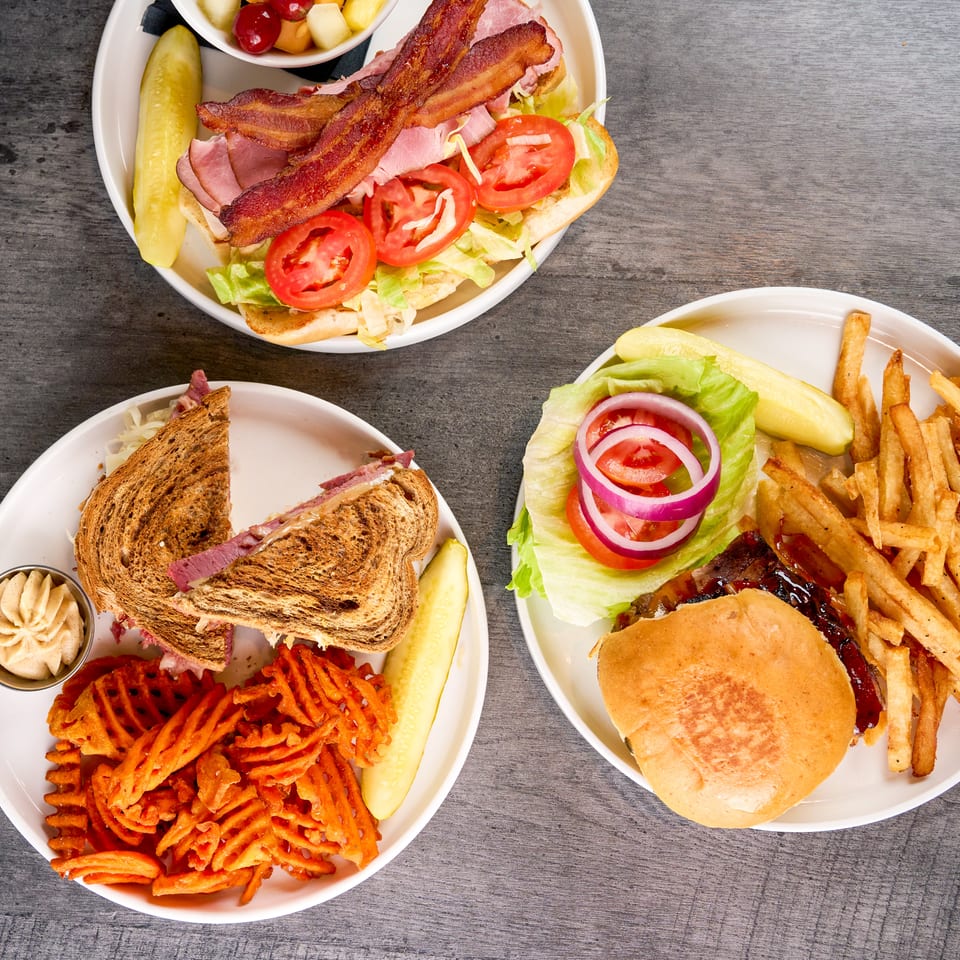Overhead view of three plates with a bacon sub, a Reuben sandwich, and a BBQ burger with fries.