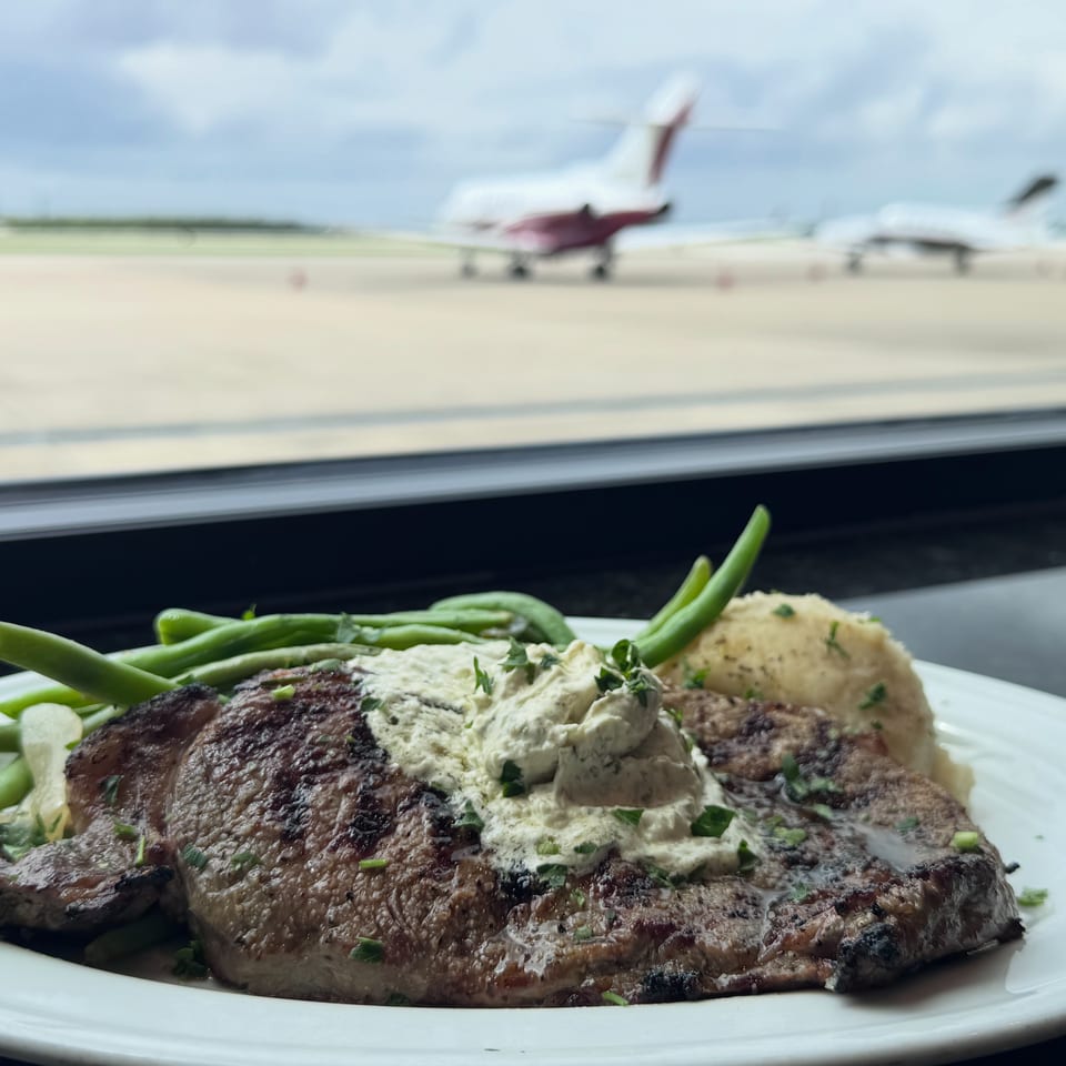 Grilled steak topped with herb butter, served with green beans and a baked potato beside an airport runway.