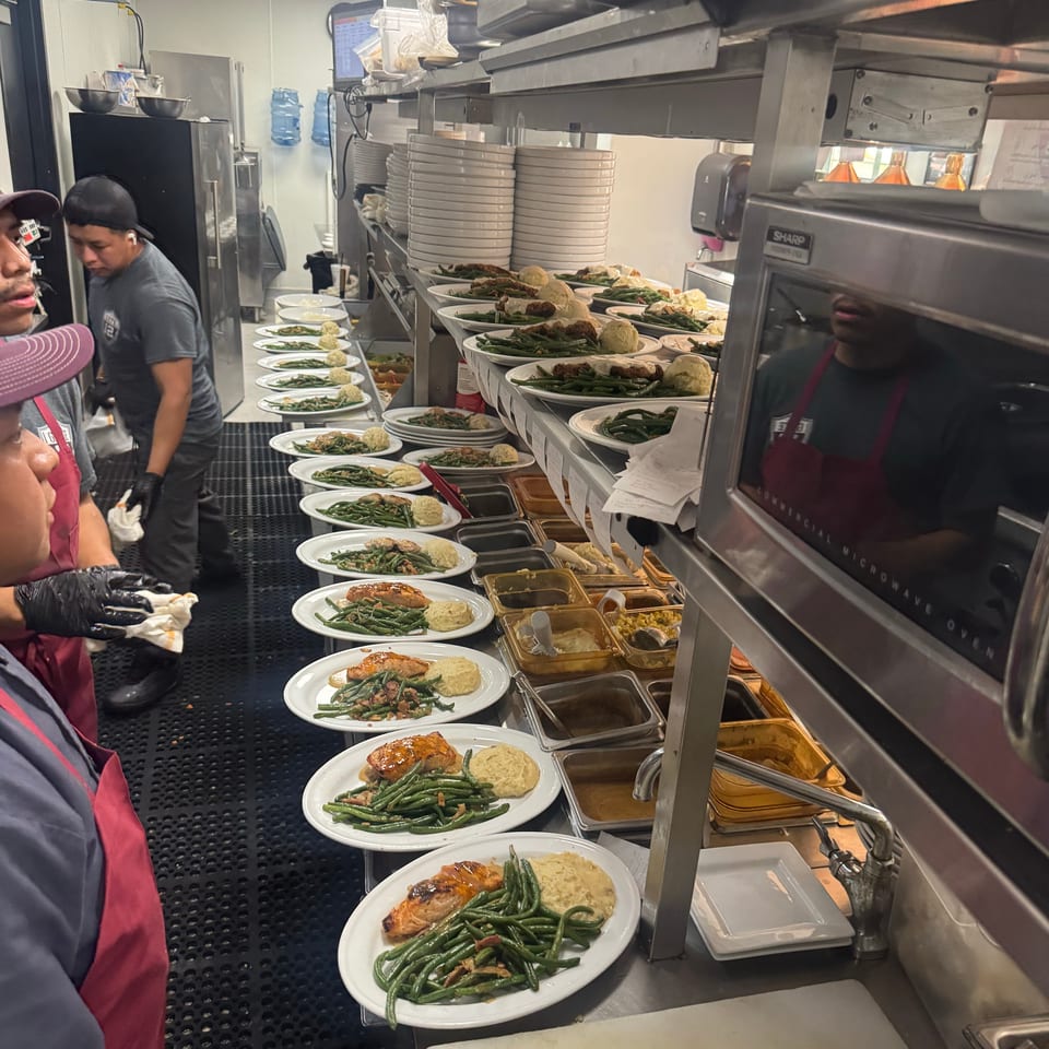Restaurant kitchen staff prepare plates of salmon, green beans, and mashed potatoes along a busy service counter.