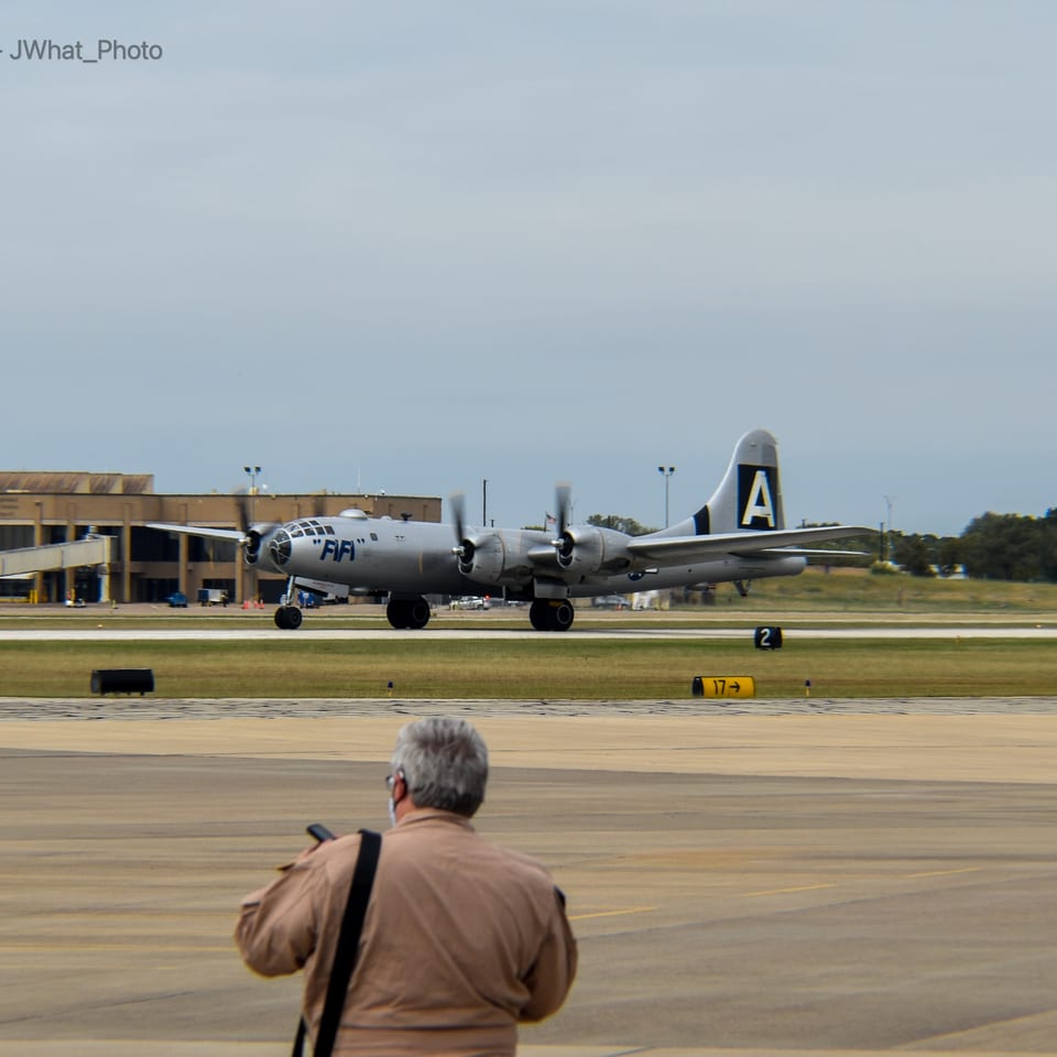 Vintage four-engine bomber taxis past an airport terminal while a spectator watches from the foreground.