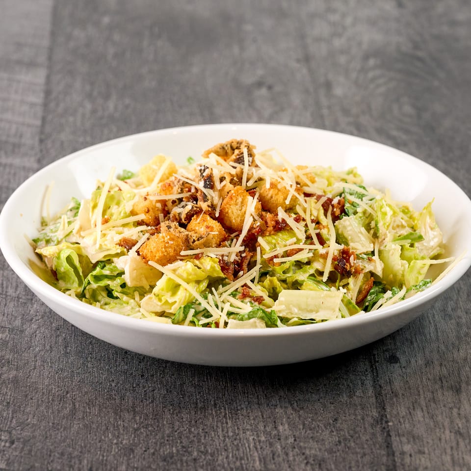 A Caesar salad in a white bowl, topped with croutons, bacon bits, and shredded Parmesan cheese.