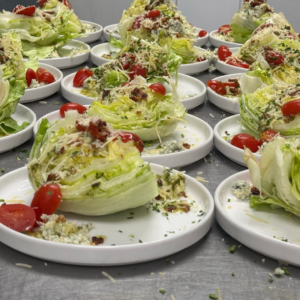 Multiple wedge salads on white plates topped with cherry tomatoes, bacon, blue cheese, herbs, and shredded cheese.