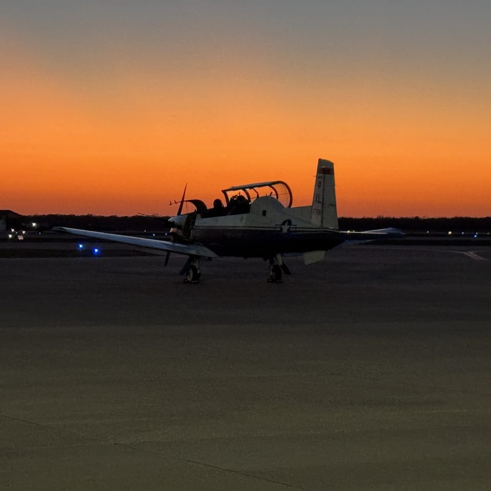 A T-6 Texan II training aircraft on the runway against a deep orange sunset.