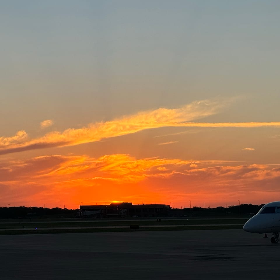 Golden sunset with glowing clouds over an airport runway, silhouetted buildings, and a private jet in the foreground.