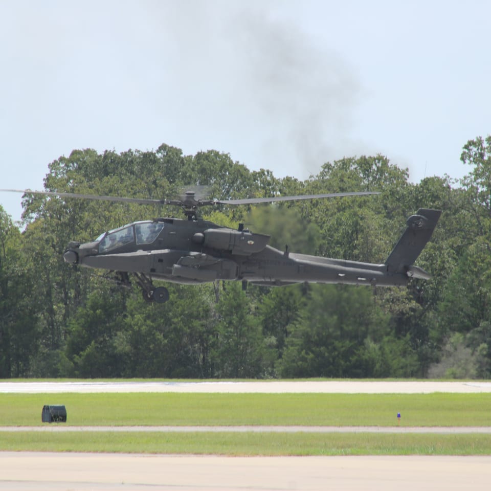 Black military attack helicopter hovering above a runway, with trees and blue sky in the background.