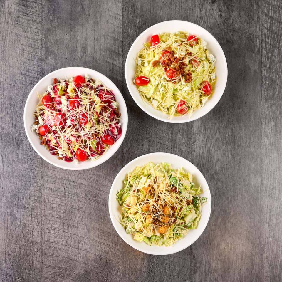Three bowls of fresh salads with various toppings like tomatoes, cheese, and bacon on a wooden table.