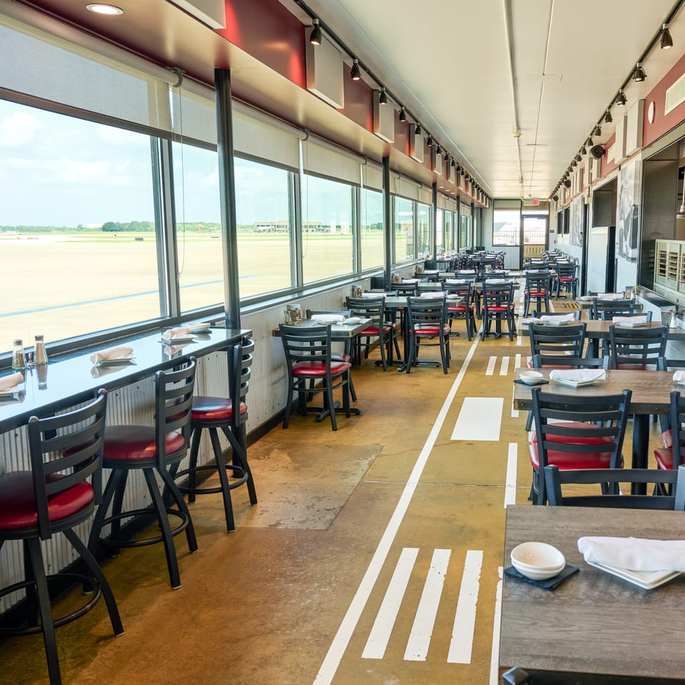 Restaurant dining room with tables and bar seating overlooking an airport runway through large windows.