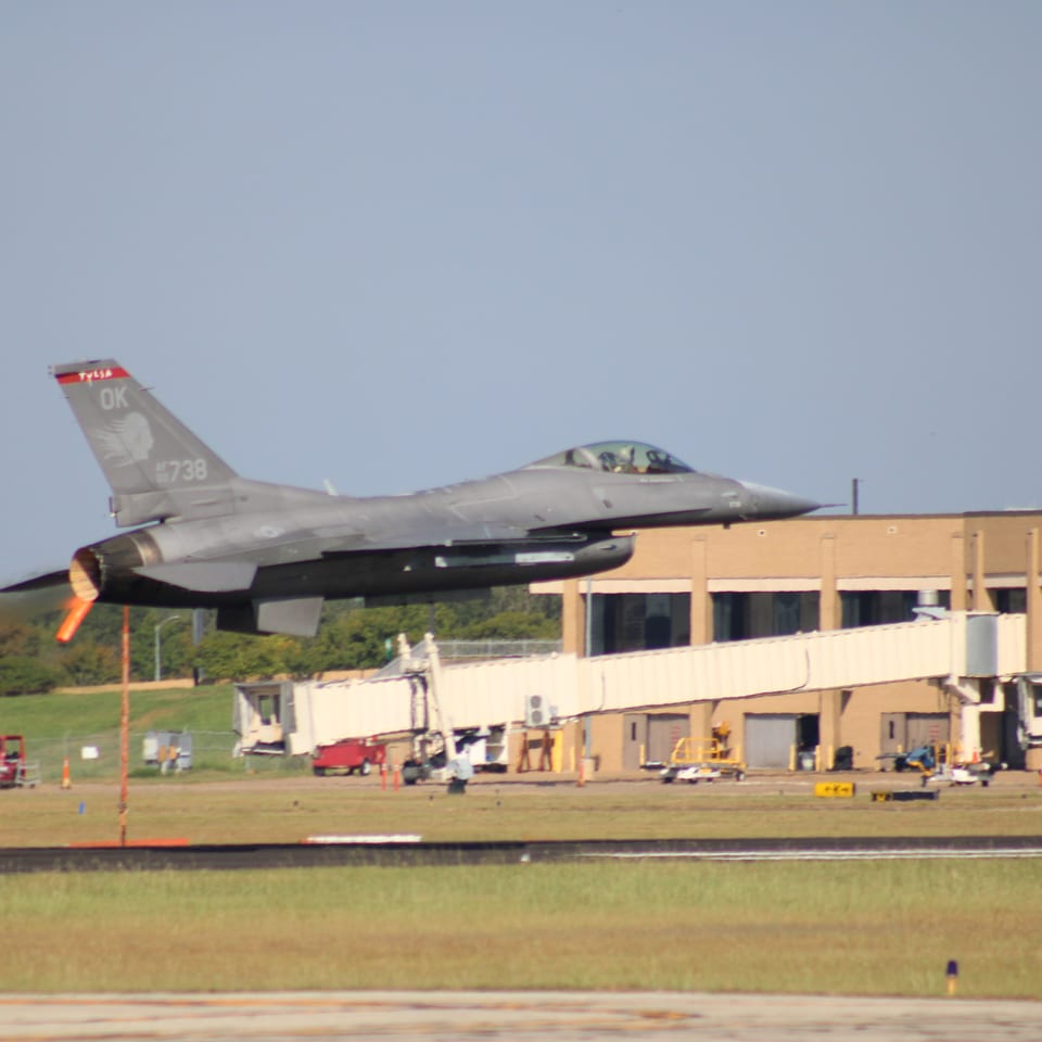 A gray fighter jet takes off above an airport runway, with fire trucks and terminal buildings in the background.