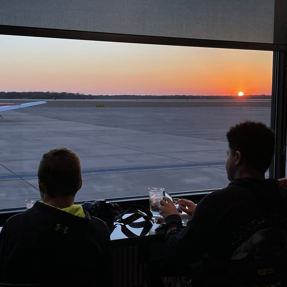 Diners sit by a restaurant window watching a small airplane on the tarmac during sunset.