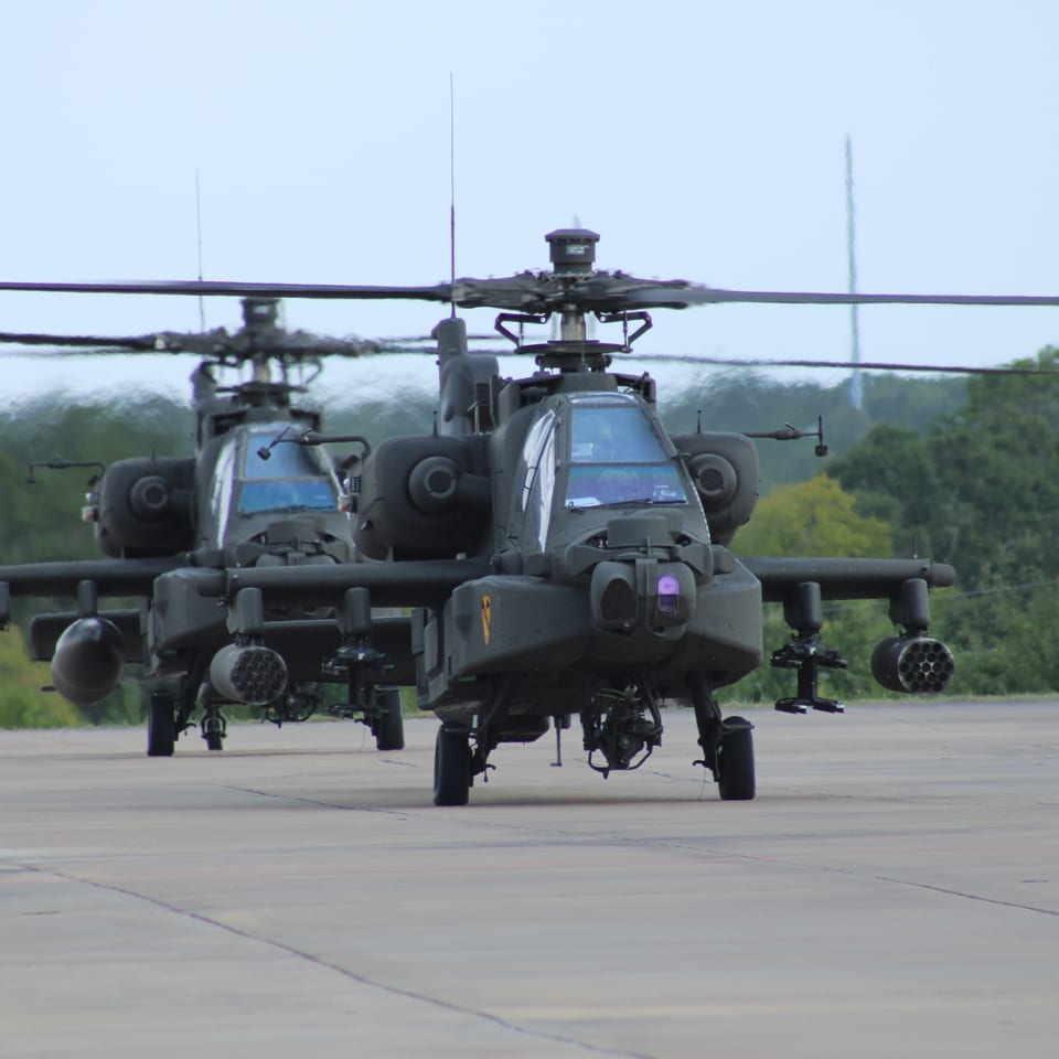 Two Apache attack helicopters parked on a runway, with forested hills in the background.