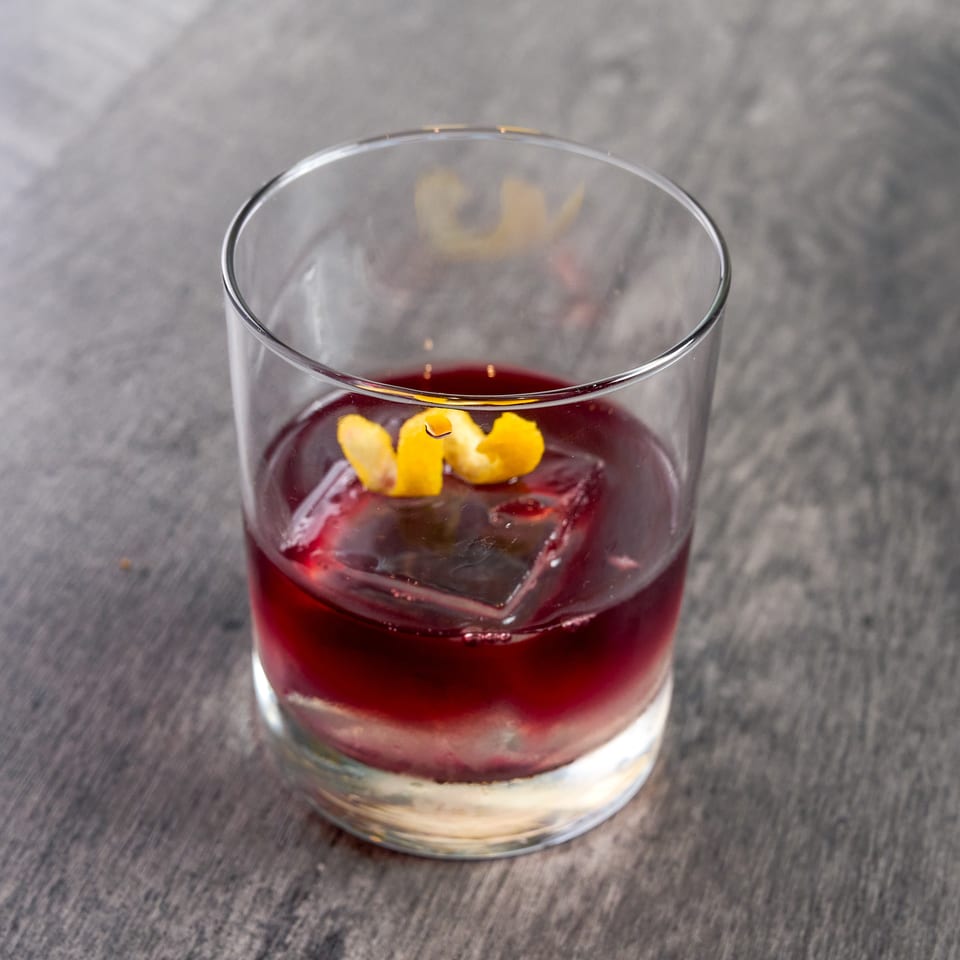 A dark red cocktail with a large ice cube and lemon peel garnish in a rocks glass.