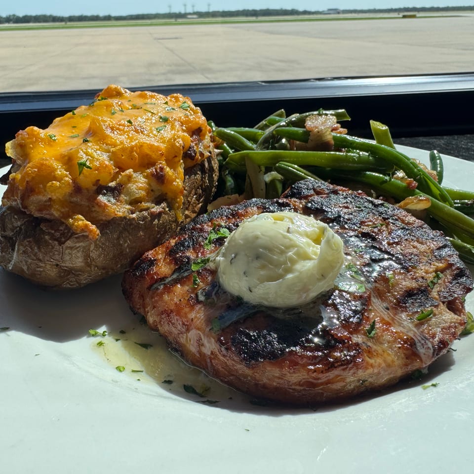 Grilled steak topped with herb butter, served with a cheese-covered baked potato and green beans on a white plate.