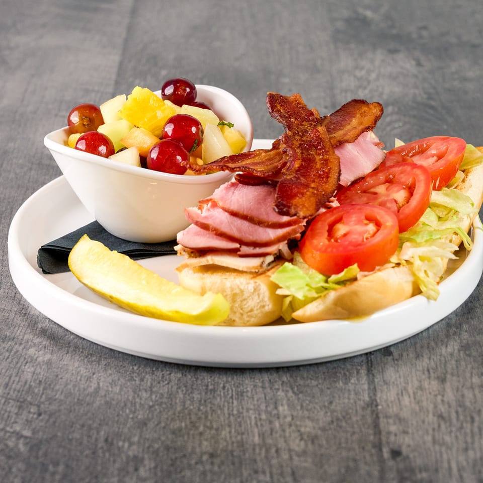 A sub sandwich with ham, bacon, lettuce, and tomato, served with a fruit cup and pickle.