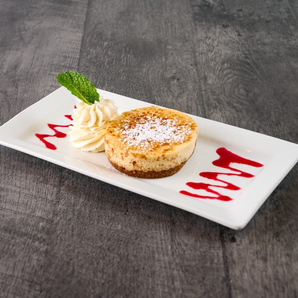 Mini cheesecake with powdered sugar, whipped cream, mint leaf, and raspberry drizzle on a white plate.