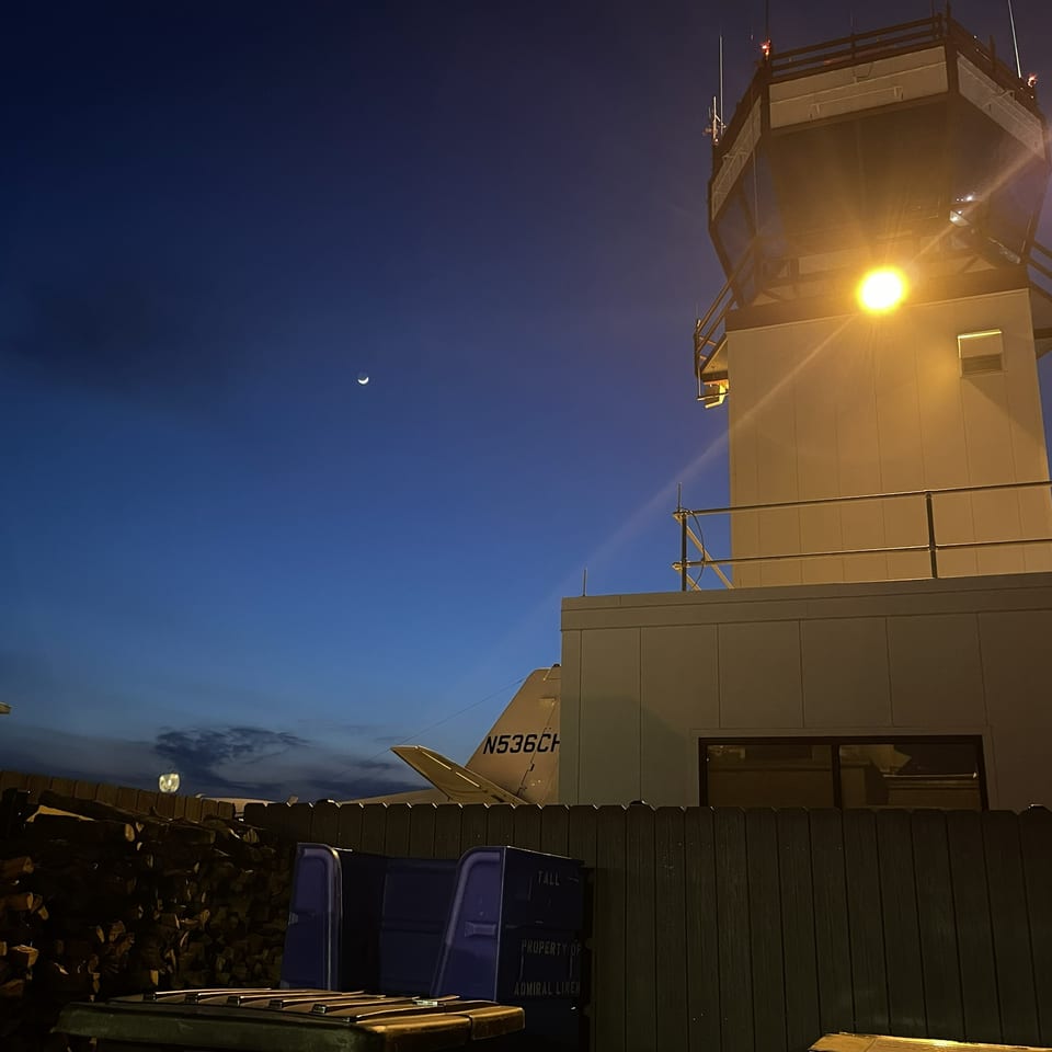 Nighttime view of a brightly lit control tower, crescent moon, rooftop railings, stacked firewood, and outdoor bins.