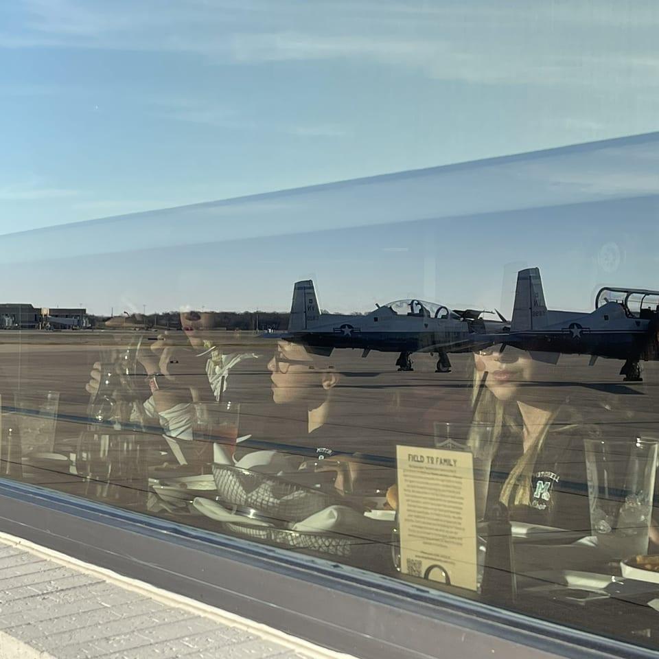 Two military trainer aircraft parked on an airfield, viewed through a window reflecting people and indoor displays.