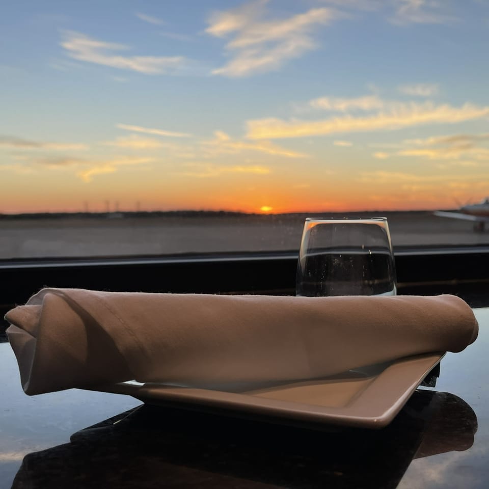 A window table setting with a glass and napkin framing a runway sunset and parked aircraft.