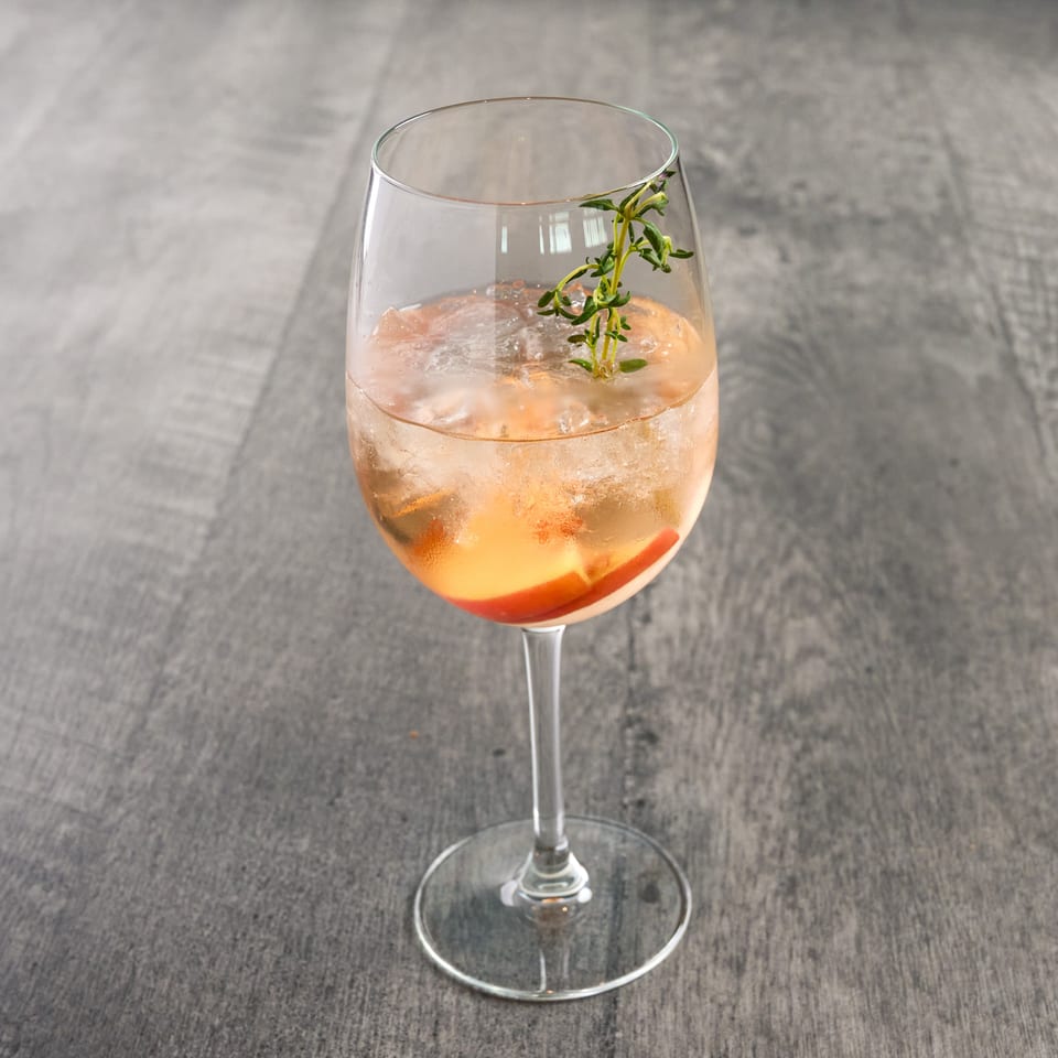 A peach-colored cocktail with ice, peach slices, and a sprig of thyme in a wine glass.