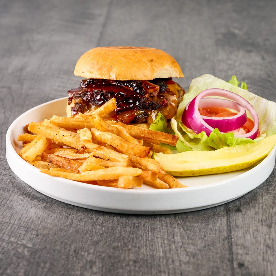 A BBQ burger with fries, lettuce, tomato, red onion, and a pickle spear on a white plate.