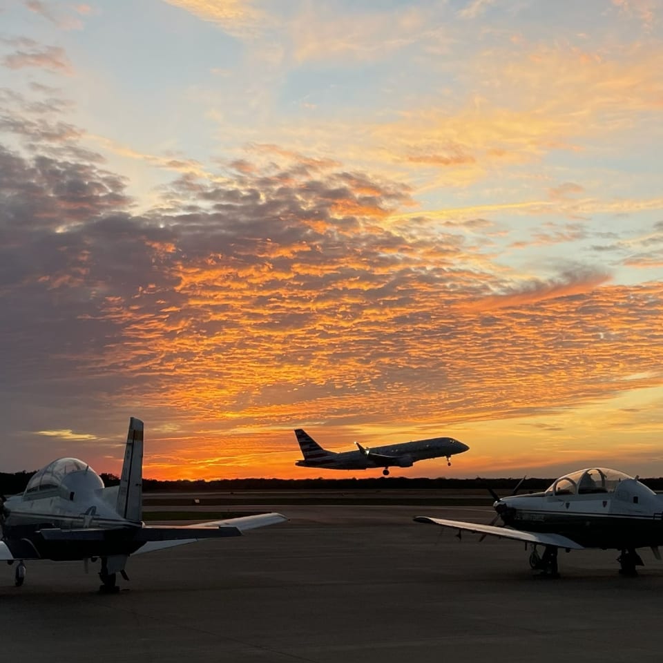 Two training jets on the tarmac as an airliner lifts off into a sunset sky.