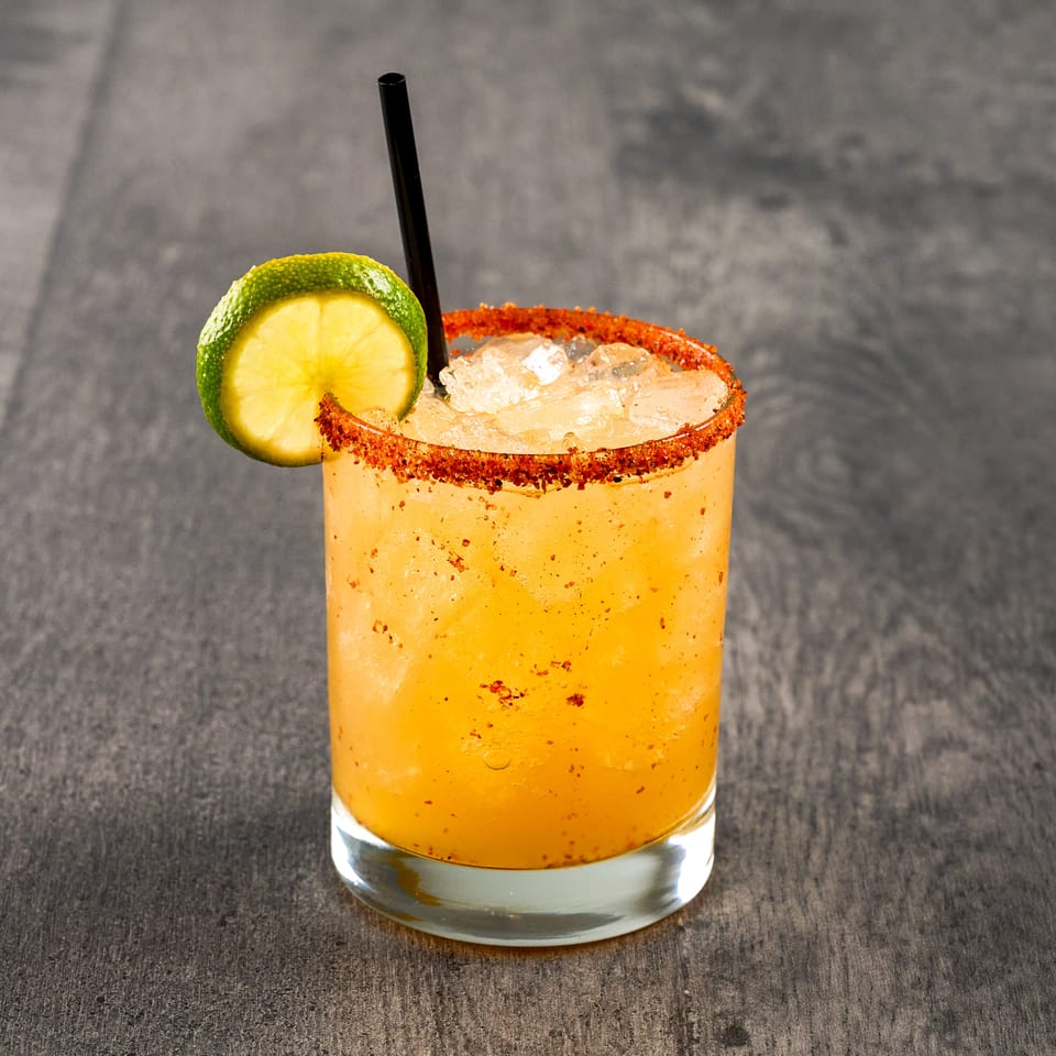 A vibrant orange cocktail with ice, a black straw, and a lime slice, garnished with chili salt.