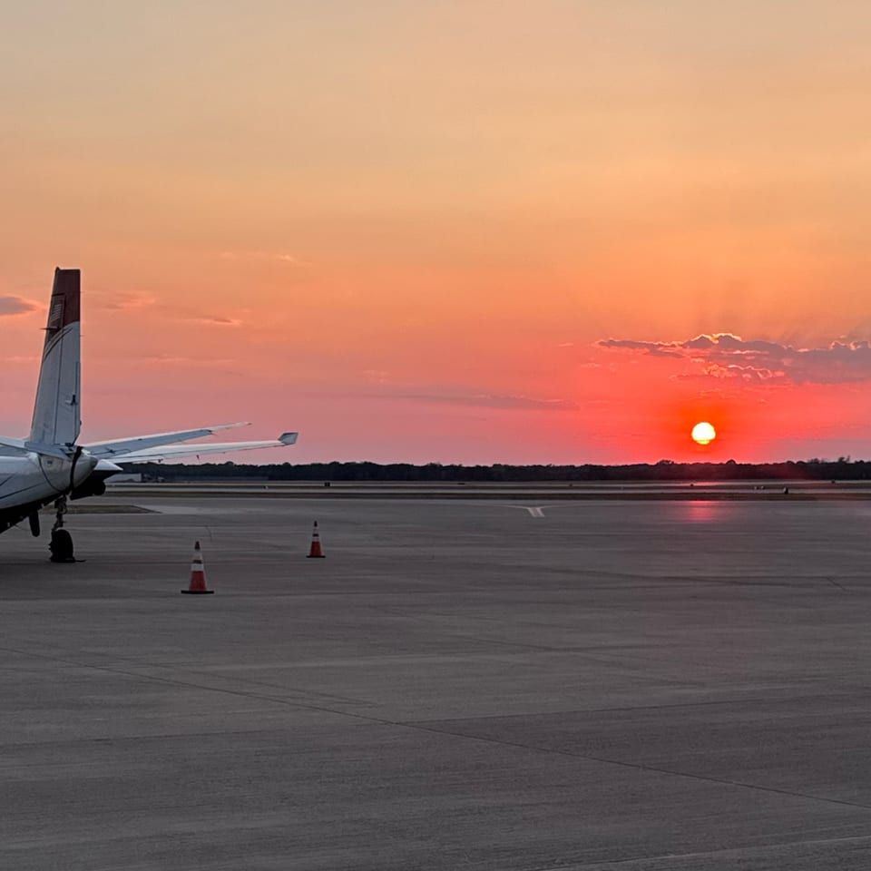 Small twin-engine airplane parked on an airport runway at sunset, with orange skies and traffic cones in the foreground.