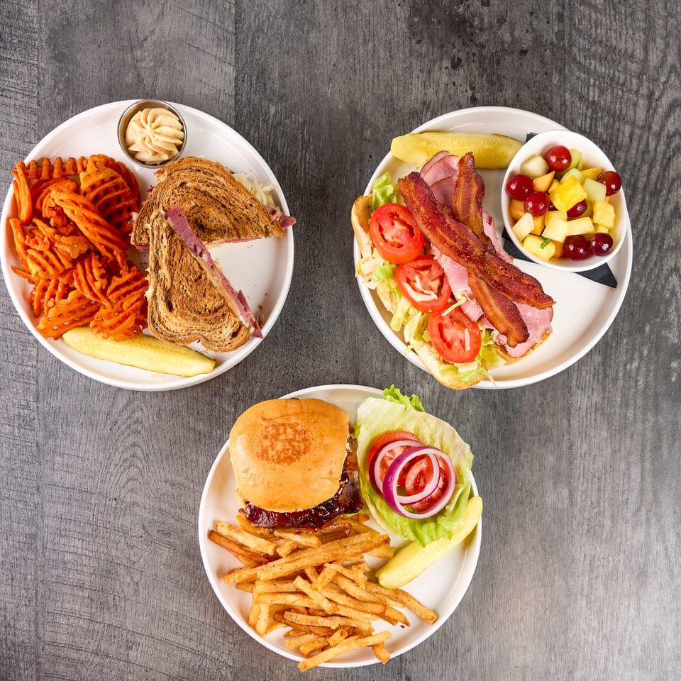 Three plates of sandwiches, fries, and fruit salad on a rustic wooden table.