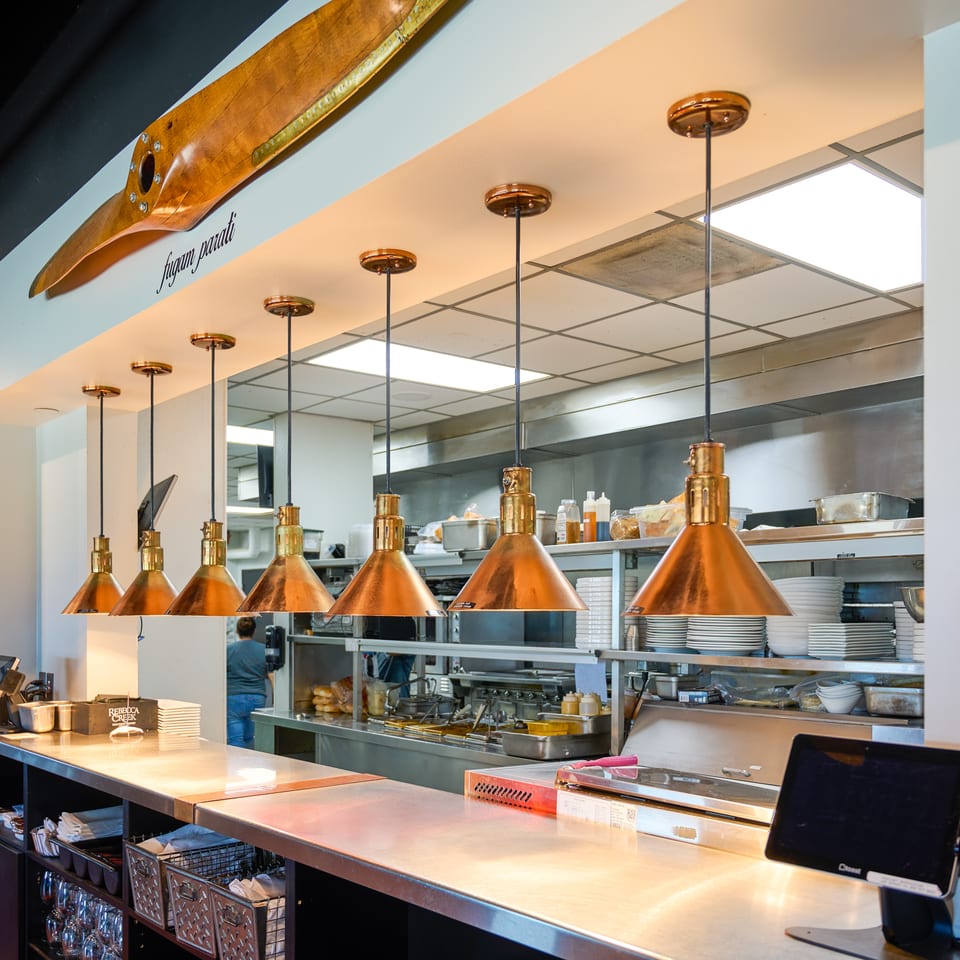 A restaurant kitchen pass-through with copper pendant lights, a wooden propeller decor, and stainless steel counters.