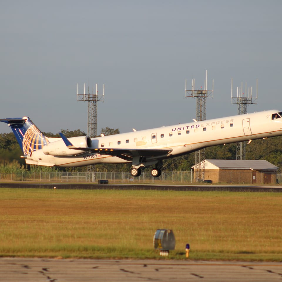 United Express regional jet landing on a runway, with airport antennas, trees, and a small utility building behind it.