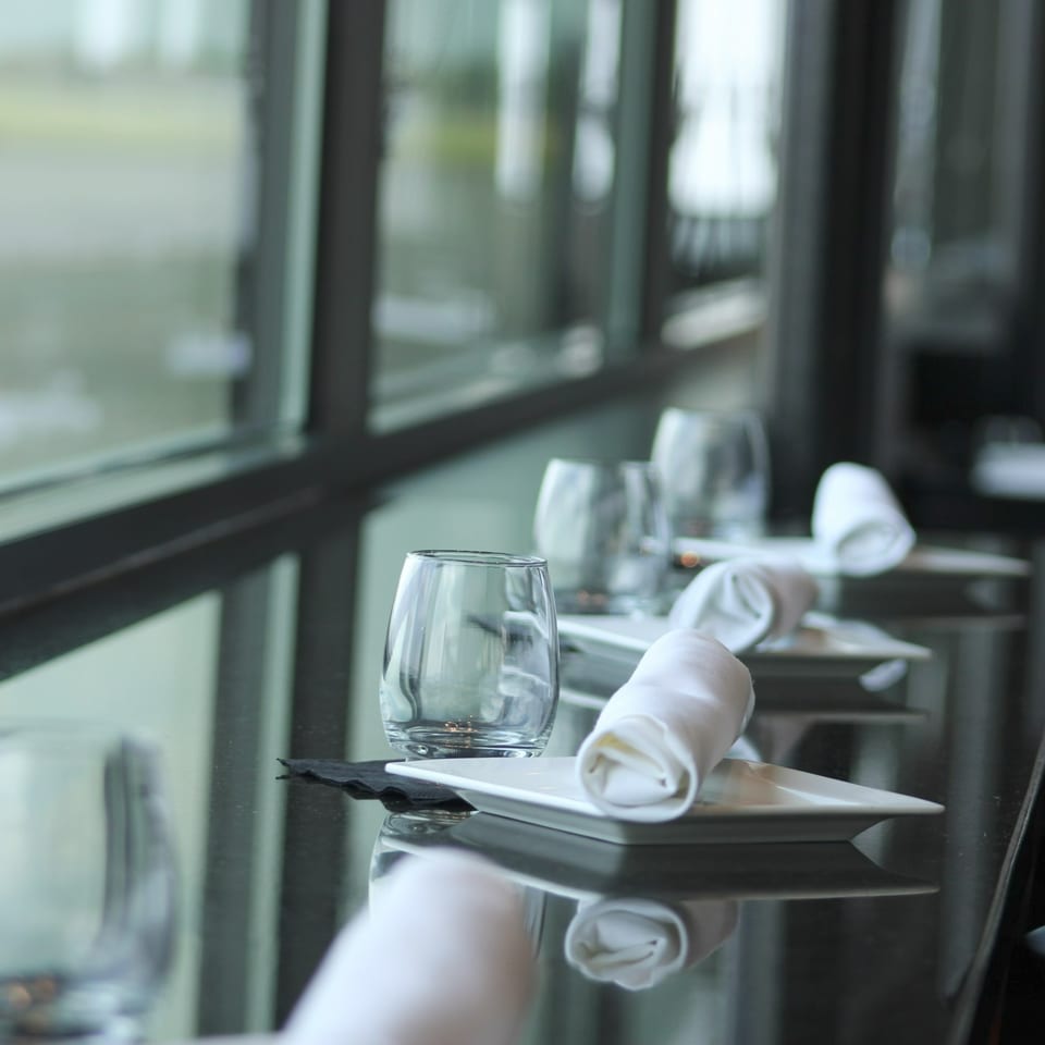 Elegant restaurant table set with white plates, rolled napkins, and wine glasses beside large windows.
