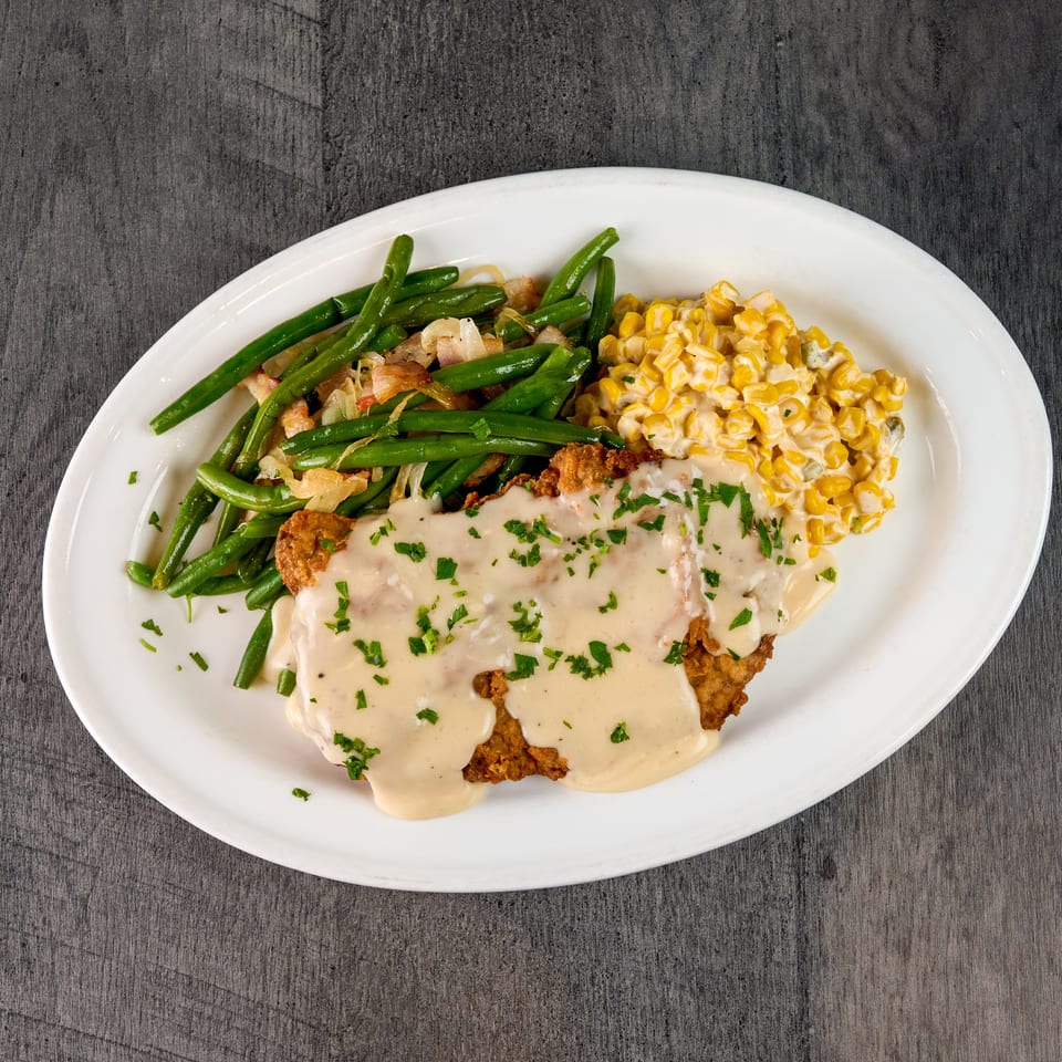 Chicken fried steak with gravy, green beans, and creamed corn on a white oval plate.