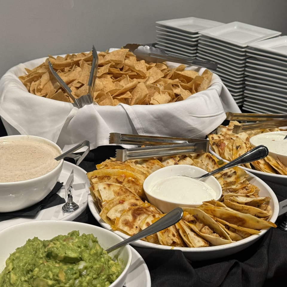 Buffet spread with tortilla chips, chicken quesadillas, guacamole, creamy dips, serving tongs, and stacked plates.
