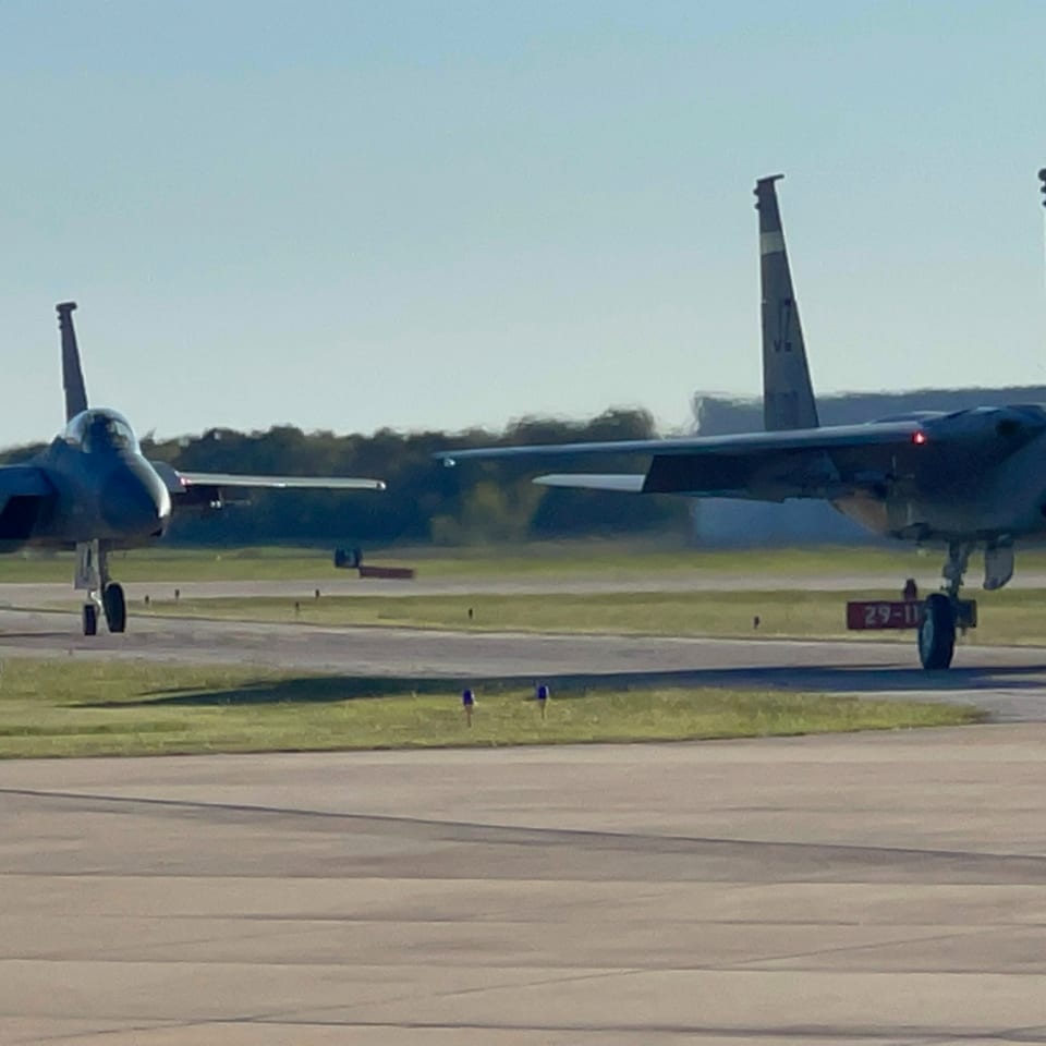 Two military fighter jets taxi on an airfield runway, with landing gear down and a tree-lined horizon.