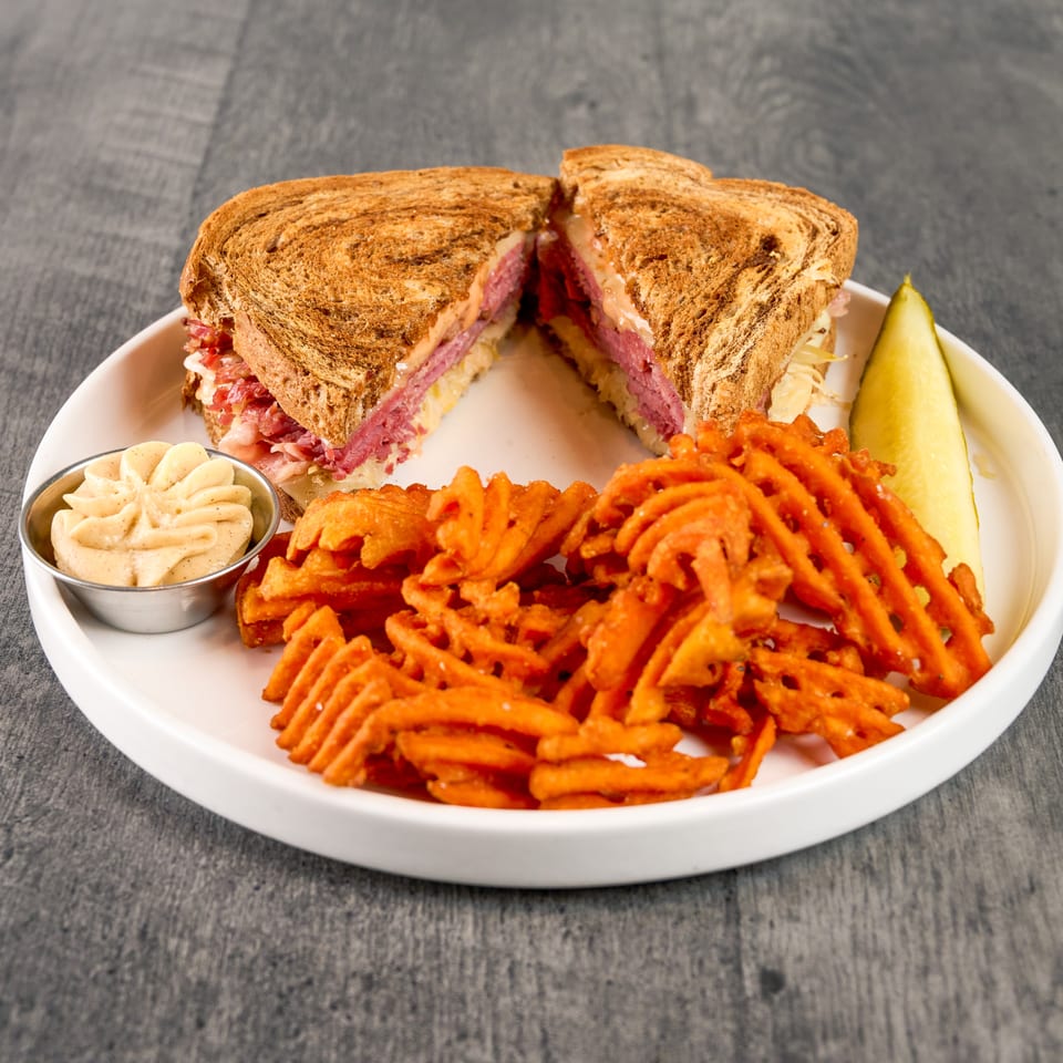 Reuben sandwich cut in half, with a side of waffle fries, a pickle spear, and dipping sauce.