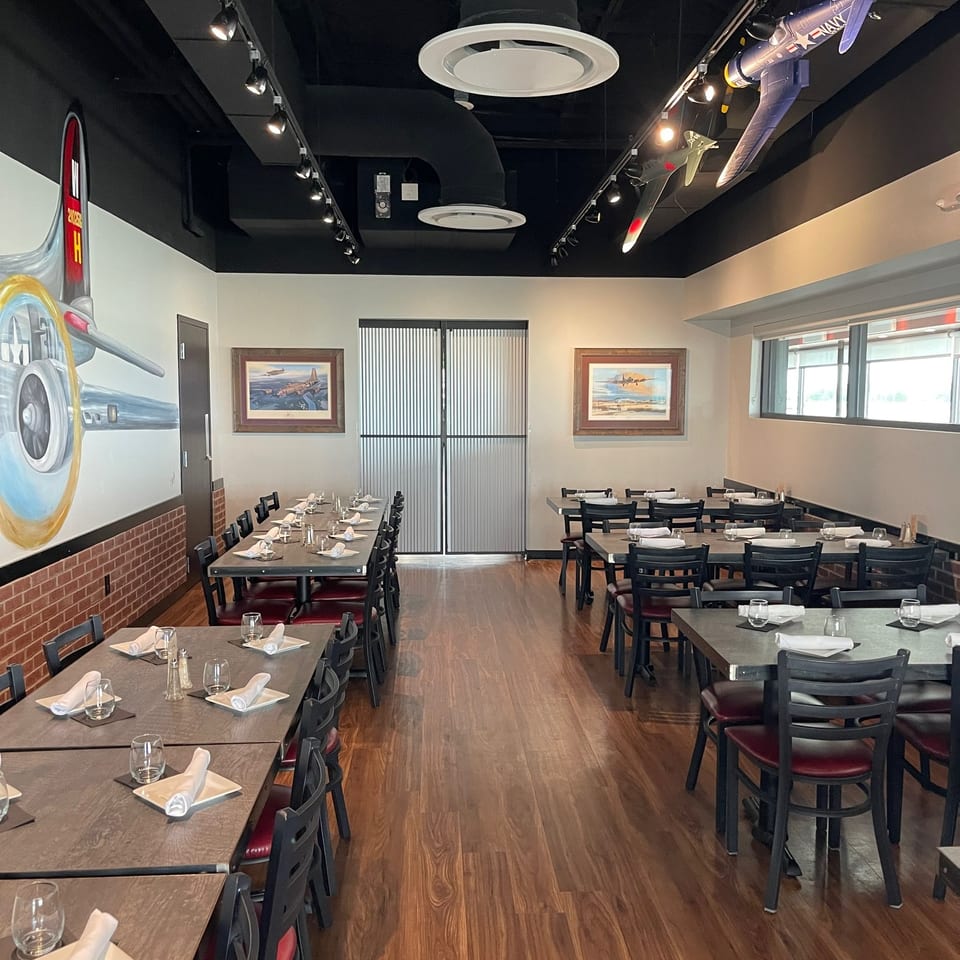 Aircraft-themed restaurant dining room with set tables, black chairs, aviation artwork, airplane models, and wood flooring.