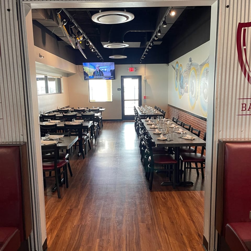 Restaurant dining room with neatly set tables, dark chairs, wood flooring, mural artwork, and wall-mounted television.