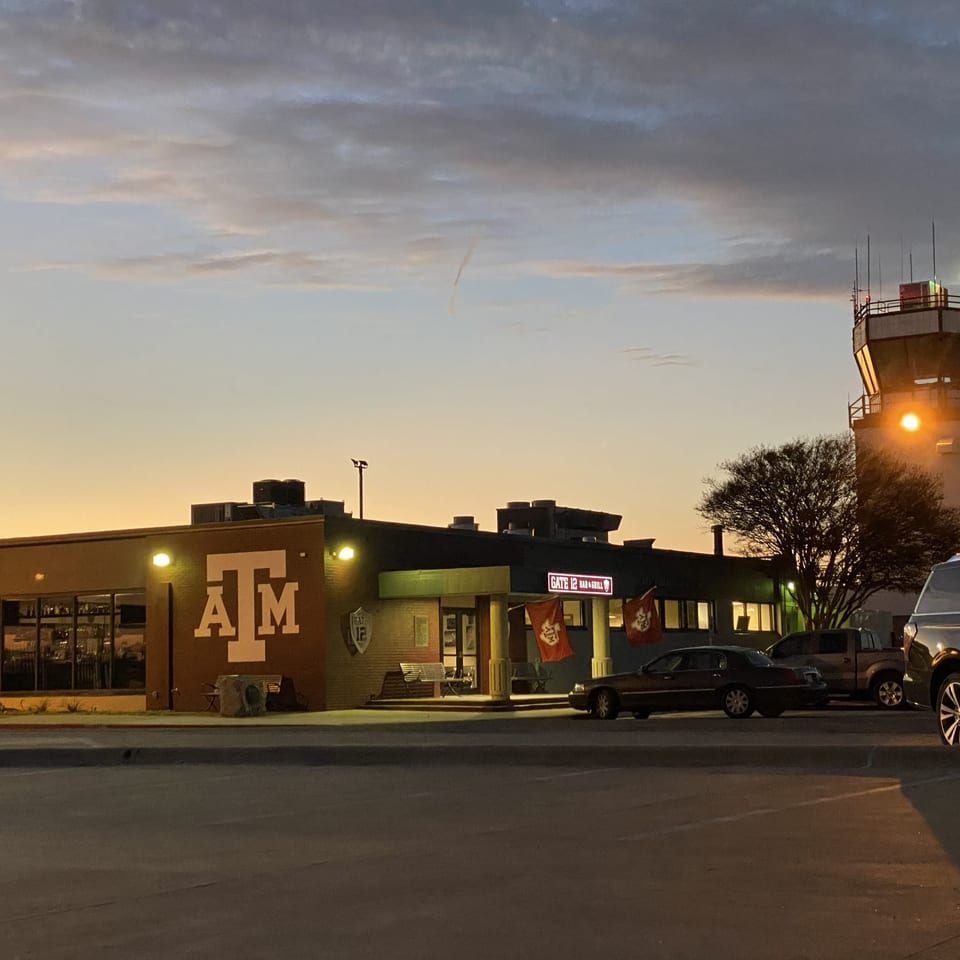 Gate 16 Bar & Grill exterior at dusk, featuring Texas A&M branding, parked cars, and an illuminated airport control tower.