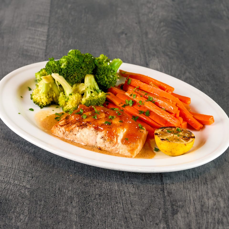 Salmon fillet with sauce, broccoli, carrots, and a grilled lemon half on a white plate.
